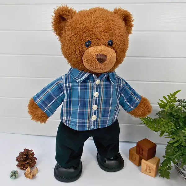 Teddy bear wearing a blue checked shirt, black trousers & black shoes. The teddy bear is standing in front of a white background with a green fern plant to the right. There are 3 wooden blocks with the letters, B,D and B on the floor 