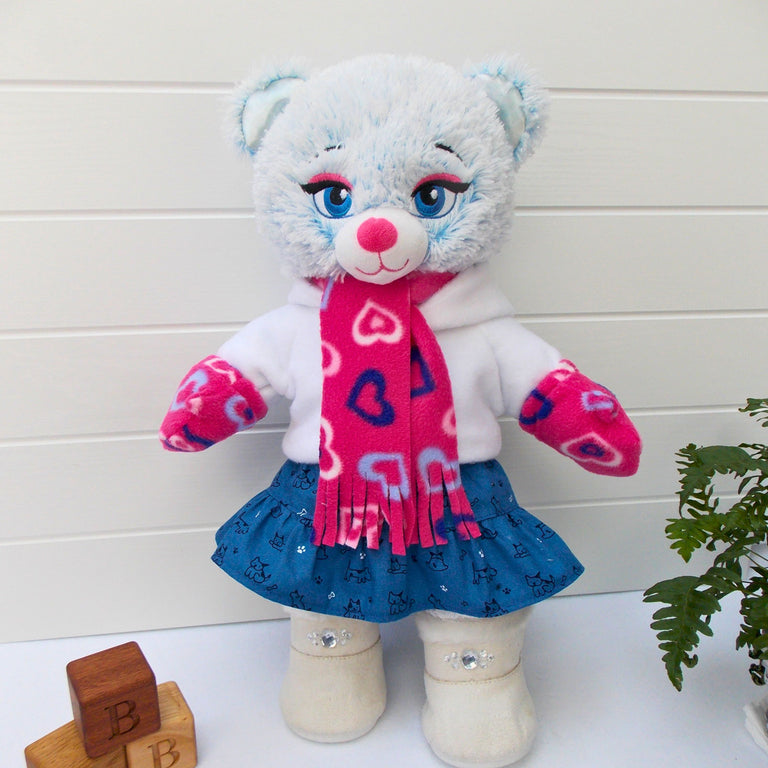 Build a bear teddy bear wearing a blue patterned skirt, white hoodie top, pink patterned scarf and mittens, and white coloured boots. The teddy bear is standing in front of a white background with a green fern plant to the right. There are some wooden bricks to the bottom left hand corner of the picture. The teddy bear skirt, teddy bear hoodie, teddy bear scarf and teddy bear mittens are made from sewing patterns from Best Dressed Bears.