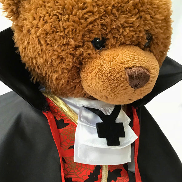 Build a bear teddy bear wearing a Halloween costume comprising of a long black cloak, red and black waistcoat with a bat pattern and gold trim, white neck tie and a black cross ‘necklace’. The teddy bear is standing in front of a white background. The teddy bear Halloween costume has been made from a sewing pattern by Best Dressed Bears.