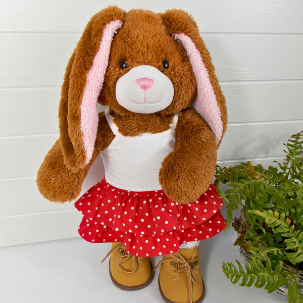Build a bear teddy bear wearing a red and white spotted skirt, white camisole top and sand coloured boots. The teddy bear is standing in front of a white background with a green fern plant to the right. The teddy bear skirt and teddy bear top are made from sewing patterns from Best Dressed Bears.