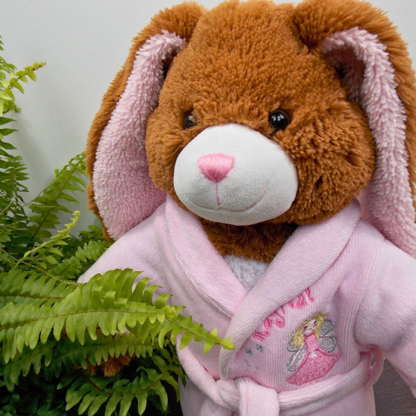 Brown teddy bear with long ears wearing a pink dressing gown with a fairy motif. The teddy bear is standing in front of a white background with a green fern plant to the left.