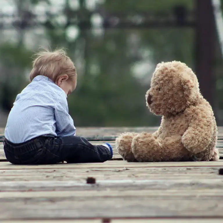 Teddy bear and child sitting on wooden floor. The child has brown hair & is wearing a blue striped shirt and black trousers. The teddy bear is not wearing any clothes. The teddy bear is watching the child