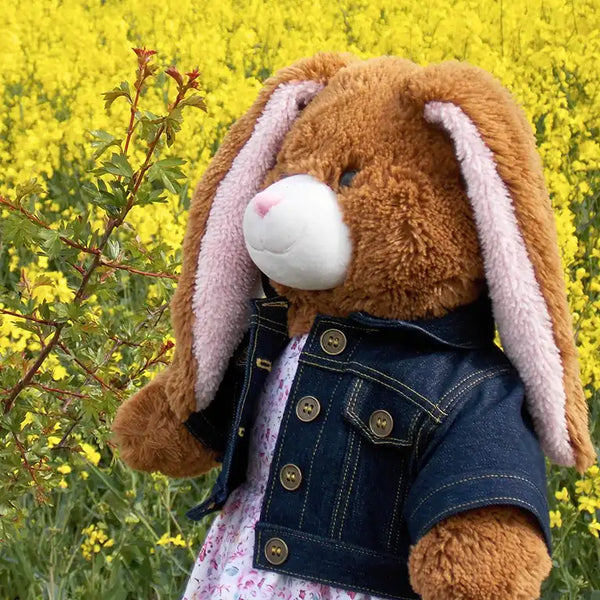 Build a bear teddy bear wearing a blue denim jacket and pink and white patterned dress standing in front of a green bush with yellow flowers
