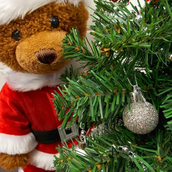 Build a bear teddy bear wearing a Santa Claus outfit standing behind a Christmas Tree