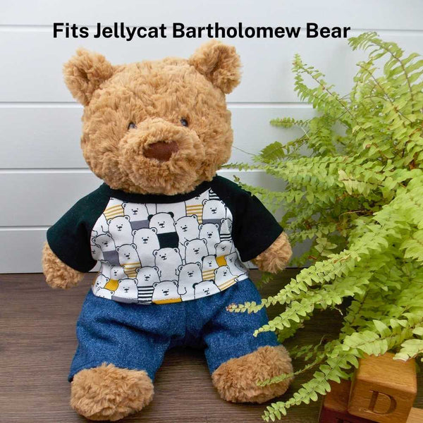 Jellycat Bartholomew Bear wearing a black patterned t-shirt and blue denim trousers. The Jellycat bear is sitting on a wooden surface with a green fern plant to the right