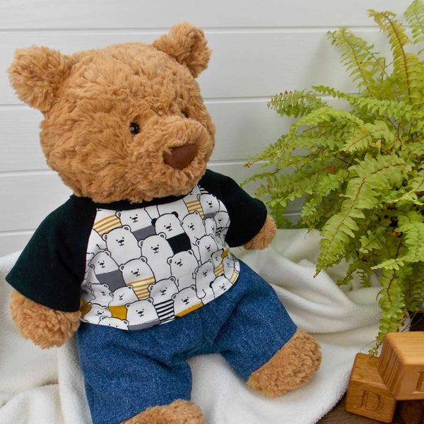 Jellycat Bartholomew Bear wearing a black patterned t-shirt and blue denim trousers. The Jellycat bear is sitting on a white blanket with a green fern plant to the right