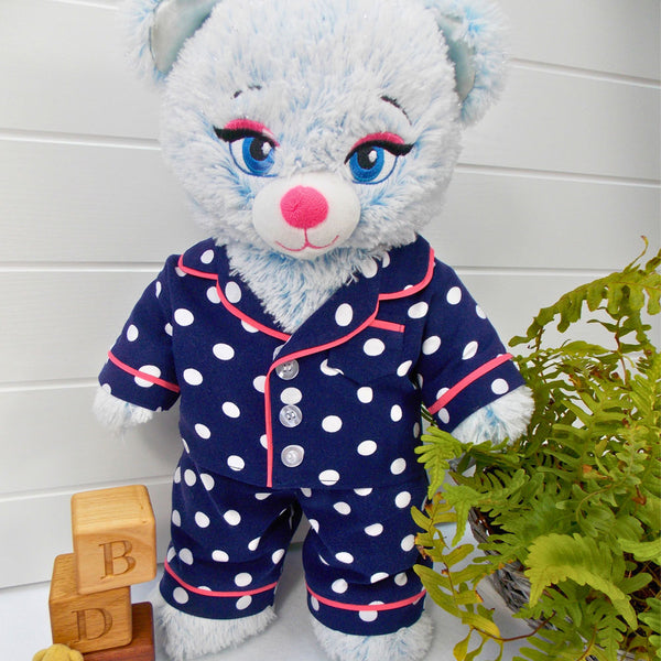 Build a bear teddy bear wearing a blue spotty pajamas with a pink trim. The teddy bear is standing in front of a white background with a green fern plant to the right. The teddy bear pyjamas were made from a sewing pattern by Best Dressed Bears.