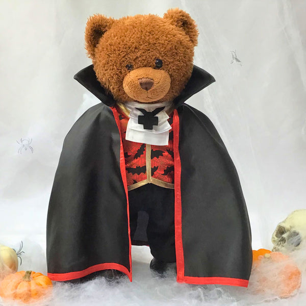 Build a bear teddy bear wearing a Halloween costume comprising of a long black cloak, red and black waistcoat with a bat pattern and gold trim, white neck tie with black cross ‘necklace’, black trousers, and black shoes. The teddy bear is standing in front of a white background. There is a skull and three pumpkins on the floor. The teddy bear Halloween costume has been made from a sewing pattern by Best Dressed Bears.