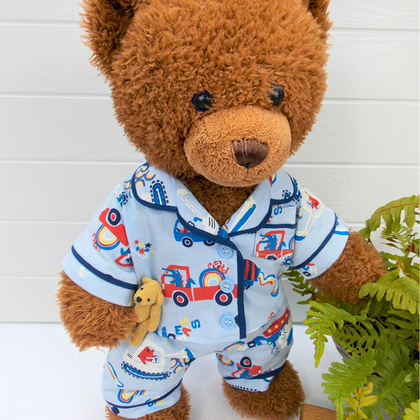 Build a bear teddy bear wearing a blue patterned pajamas with a dark blue trim. The teddy bear is holding a little beige teddy bear and is standing in front of a white background with a green fern plant to the right. The teddy bear pyjamas were made from a sewing pattern by Best Dressed Bears.