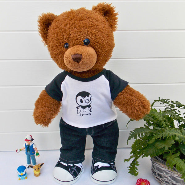 A brown teddy bear wearing a white and black t-shirt with a Pokemon character on the front, black denim pants, and black and white trainers. The teddy bear is standing in front of a white background with a green fern plant to the right. There are various Pokemon figures on the floor. The teddy bear t-shirt & teddy bear pants were made from sewing patterns by Best Dressed Bears.