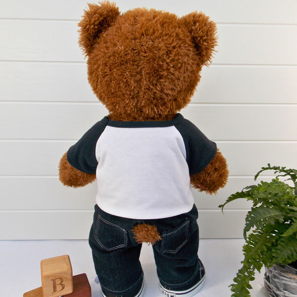 Build a bear teddy bear wearing a white and black t-shirt, black denim pants, and black and white trainers. The teddy bear is standing in front of a white background with a green fern plant to the right. The teddy bear t-shirt & teddy bear pants were made from sewing patterns by Best Dressed Bears.