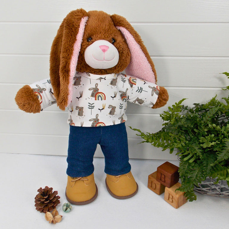 Build a bear teddy bear wearing a white patterned t-shirt, blue denim pants and sand coloured boots. The teddy bear is standing in front of a white background with a green fern plant to the right. The teddy bear t-shirt & teddy bear pants were made from sewing patterns by Best Dressed Bears.