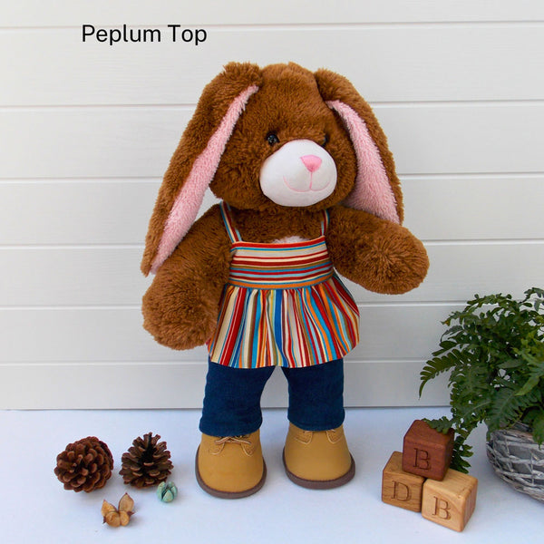 Build a bear teddy bear wearing a striped peplum top, blue denim leggings & sand coloured boots. The teddy bear is standing in front of a white background with a green fern plant to the right. There are 3 wooden bricks on the floor with the letters B, D and B on them. The teddy bear t-shirt & teddy bear pants were made from sewing patterns by Best Dressed Bears.