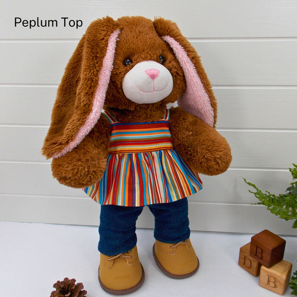 Build a bear teddy bear wearing a striped peplum top, blue denim leggings & sand coloured boots. The teddy bear is standing in front of a white background with a green fern plant to the right. There are 3 wooden bricks on the floor with the letters B, D and B on them. The teddy bear t-shirt & teddy bear pants were made from sewing patterns by Best Dressed Bears.