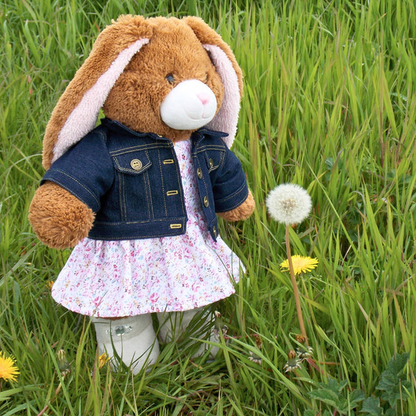 Build a bear teddy bear wearing a blue denim jacket, pink and white patterned dress and white boots. The teddy bear is standing in a field of long grass by a dandelion. The teddy bear denim jacket and teddy bear dress have been made from sewing patterns by Best Dressed Bears.