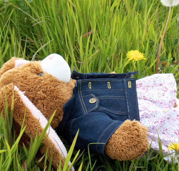 Build a bear teddy bear wearing a blue denim jacket, and a pink and white patterned dress. The teddy bear is lying in a field of long grass by a dandelion. The teddy bear denim jacket and teddy bear dress have been made from sewing patterns by Best Dressed Bears.