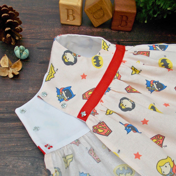 Teddy bear dress with superhero pattern and red waistband lying on a wooden surface. There are 3 wooden blocks with the initials BDB depicting the company name of Best Dressed Bears. The teddy bear dress is made from a sewing pattern by Best Dressed Bears