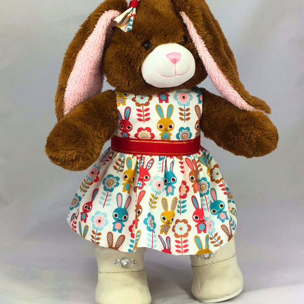 Build a bear teddy bear wearing a white dress with a bunny rabbit pattern and red ribbon waistband with white boots. The teddy bear is standing in front of a white background. The teddy bear dress is made from a sewing pattern by Best Dressed Bears