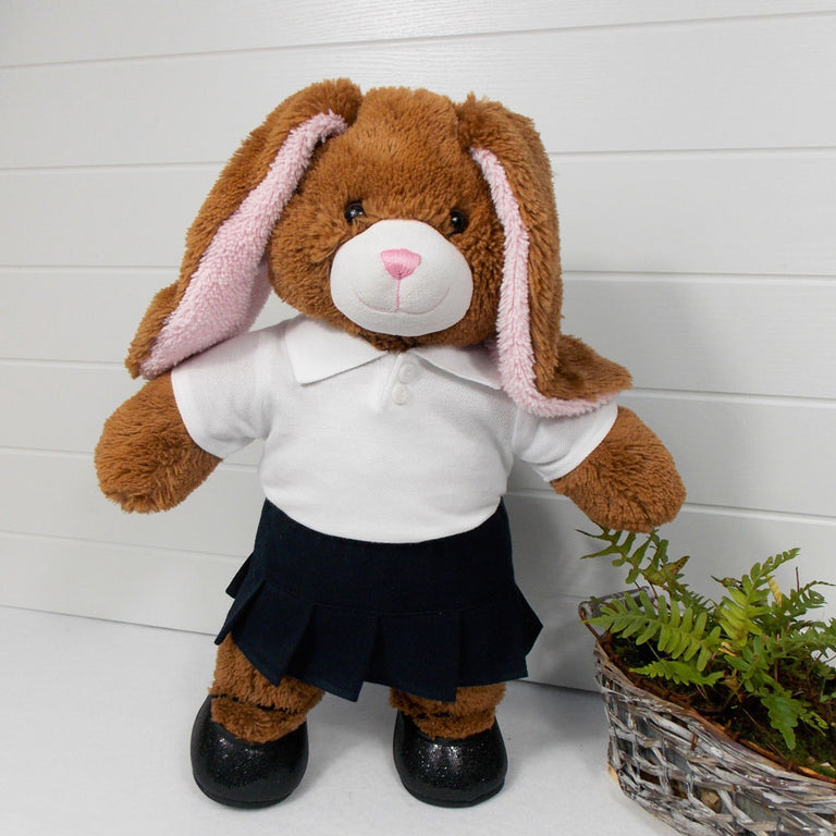 Build a bear teddy bear wearing a white polo t-shirt, navy blue pleated skirt, and black shoes. The teddy bear is standing in front of a white background with a green fern plant on the right. The teddy bear t-shirt and teddy bear skirt have been made from sewing patterns by Best Dressed Bears.
