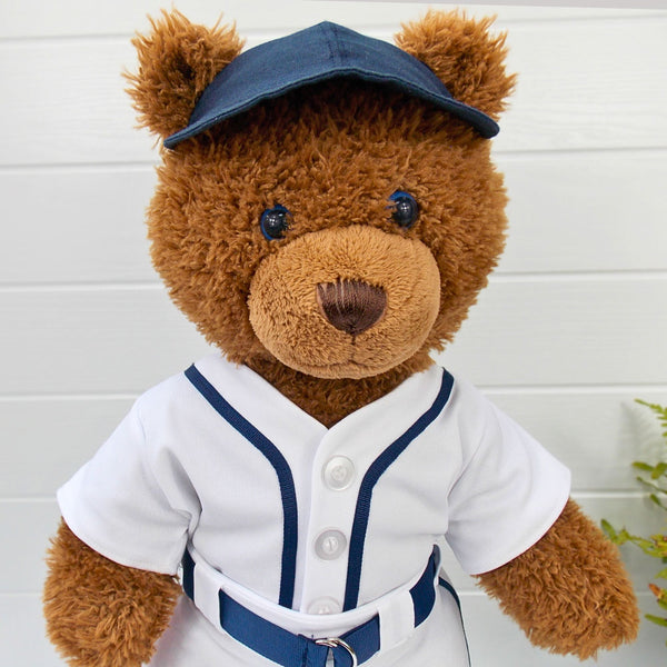 Build a bear teddy bear wearing a blue baseball cap and a white and blue baseball uniform. The teddy bear is standing in front of a white background with a small fern plant on the right. The teddy bear baseball and teddy bear baseball uniform are made from sewing patterns by Best Dressed Bears.