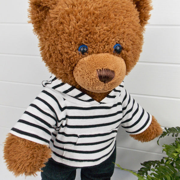 Build a bear teddy bear wearing a black and white striped hoodie and black denim jeans. The teddy bear is standing in front of a white background with a green fern plant on the right. The teddy bear hoodie and teddy bear pants have been made from sewing patterns by Best Dressed Bears.