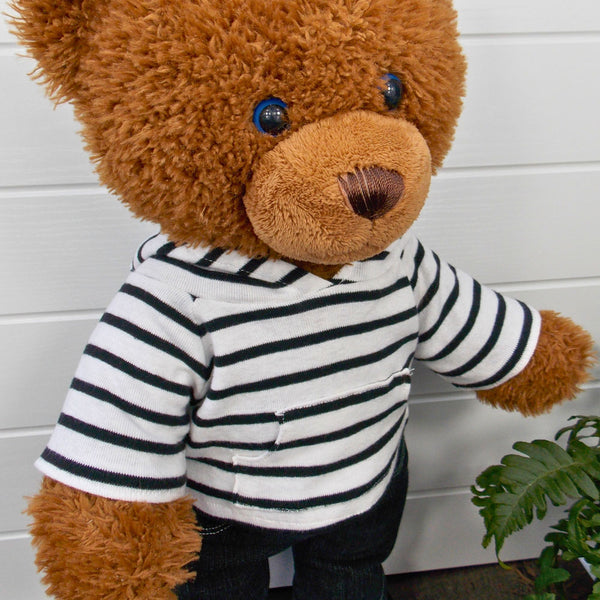 Build a bear teddy bear wearing a black and white striped hoodie and black denim jeans. The teddy bear is standing in front of a white background with a green fern plant on the right. The teddy bear hoodie and teddy bear pants have been made from sewing patterns by Best Dressed Bears.