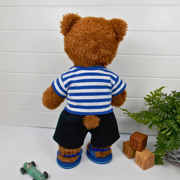 Build a bear teddy bear wearing a blue and white striped t-shirt, black shorts, and blue sandals. The teddy bear is standing in front of a white background with a green fern plant to the right. There are 3 wooden blocks with the letters B,D and B on the floor next to the fern. The teddy bear t-shirt and teddy bear shots were made from sewing patterns by Best Dressed Bears.