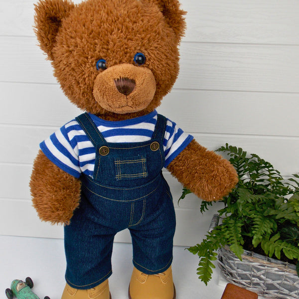 Build a bear teddy bear wearing a blue and white striped t-shirt, blue denim overalls / dungarees and sand coloured boots. The teddy bear is standing in front of a white background. The teddy bear t-shirt and teddy bear overalls / dungarees have been made from sewing patterns by Best Dressed Bears.