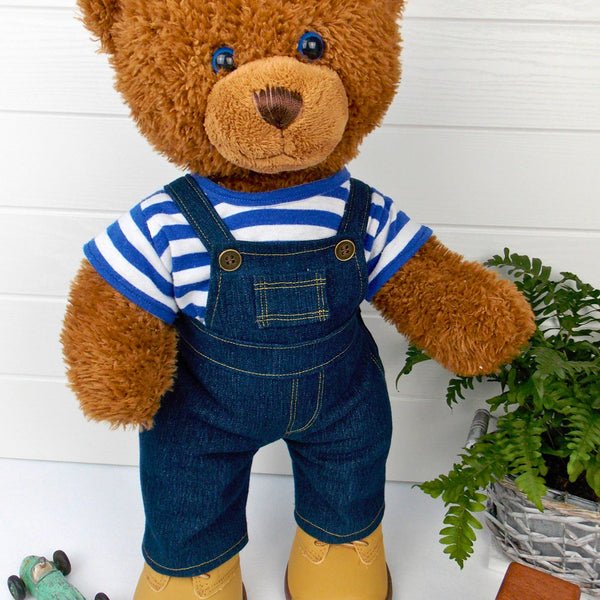Build a bear teddy bear wearing a blue and white striped t-shirt, blue denim overalls / dungarees and sand coloured boots. The teddy bear is standing in front of a white background. The teddy bear t-shirt and teddy bear overalls / dungarees have been made from sewing patterns by Best Dressed Bears.