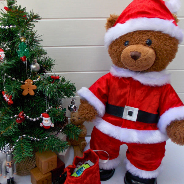 Build a bear teddy bear wearing a red and white Santa outfit. The teddy bear is standing in front of a white background and to the right of a Christmas tree. The teddy bear Santa outfit is made from a sewing pattern from Best Dressed Bears.