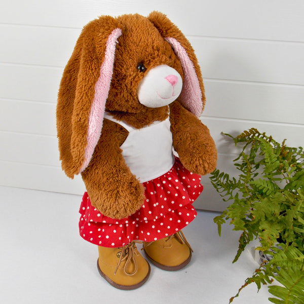 Build a bear teddy bear wearing a red and white spotted skirt, white camisole top and sand coloured boots. The teddy bear is standing in front of a white background with a green fern plant to the right. The teddy bear skirt and teddy bear top are made from sewing patterns from Best Dressed Bears.