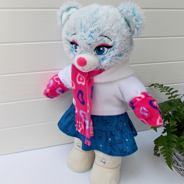 Build a bear teddy bear wearing a blue patterned skirt, white hoodie top, pink patterned scarf and mittens, and white coloured boots. The teddy bear is standing in front of a white background with a green fern plant to the right. The teddy bear skirt, teddy bear hoodie, teddy bear scarf and teddy bear mittens are made from sewing patterns from Best Dressed Bears.