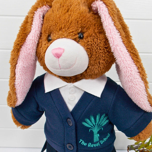 Build a bear teddy bear wearing a blue school cardigan and white polo t-shirt. The teddy bear is standing in front of a white background with a green fern plant to the right. The teddy bear school cardigan and teddy bear t-shirt are made from sewing patterns from Best Dressed Bears.
