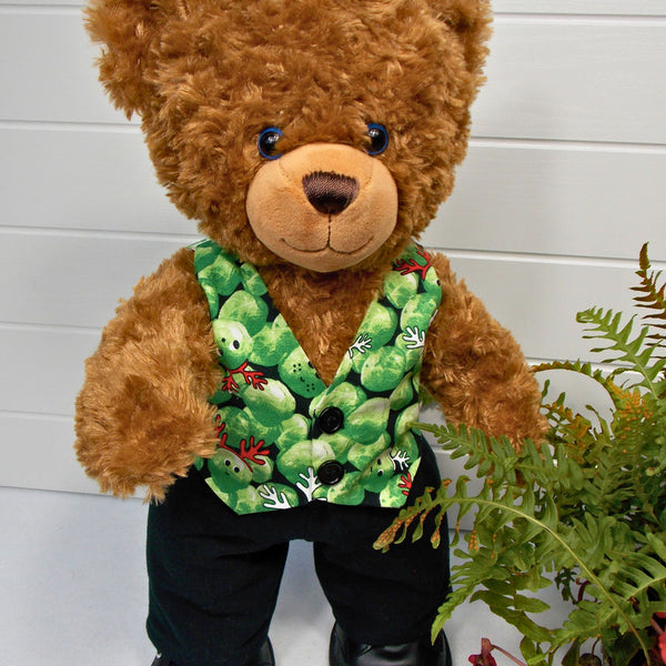 Build a bear teddy bear wearing a green patterned waistcoat, black trousers and black shoes. The teddy bear is standing in front of a white background with a green fern plant to the right. The teddy bear waistcoat and teddy bear pants were made from sewing patterns by Best Dressed Bears.