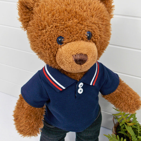 Build a bear teddy bear wearing a blue polo t-shirt and black denim jeans. The teddy bear is standing in front of a white background with a green fern plant on the right. The teddy bear t-shirt and teddy bear pants have been made from sewing patterns by Best Dressed Bears.