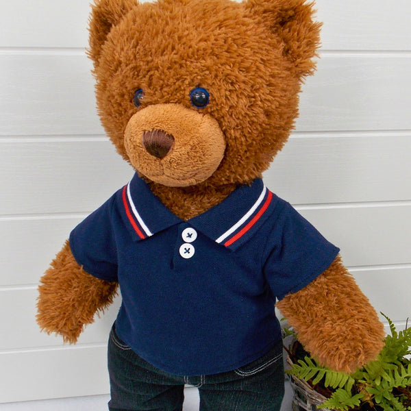 A brown coloured teddy bear wearing a blue polo t-shirt and black denim jeans. The teddy bear is standing in front of a white background with a green fern plant on the right. The teddy bear t-shirt and teddy bear pants have been made from sewing patterns by Best Dressed Bears.