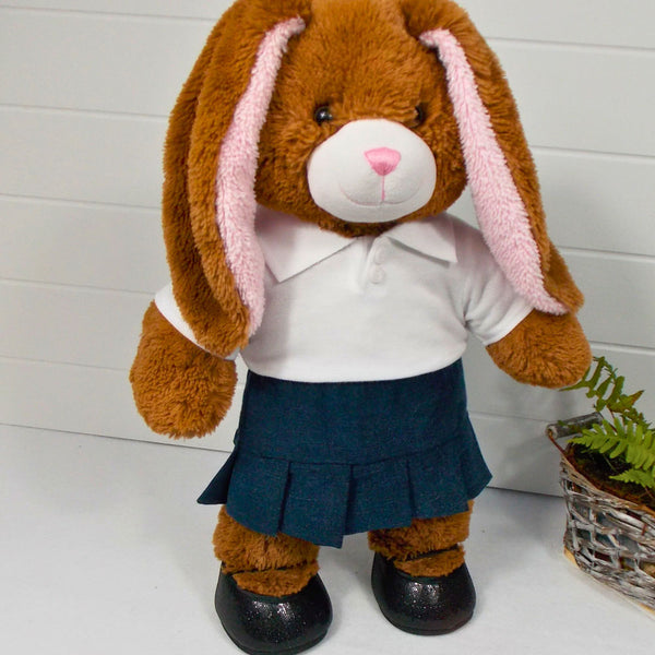 Build a bear teddy bear wearing a white polo t-shirt, navy pleated skirt, and black shoes. The teddy bear is standing in front of a white background with a green fern plant on the right. The teddy bear t-shirt and teddy bear skirt have been made from sewing patterns by Best Dressed Bears.