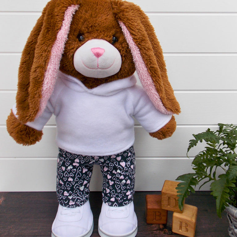 Build a bear teddy bear wearing a white hoodie, black and pink leggings and white training shoes. The teddy bear is standing in front of a white background with a green fern plant on the right. The teddy bear hoodie and teddy bear leggings have been made from sewing patterns by Best Dressed Bears.