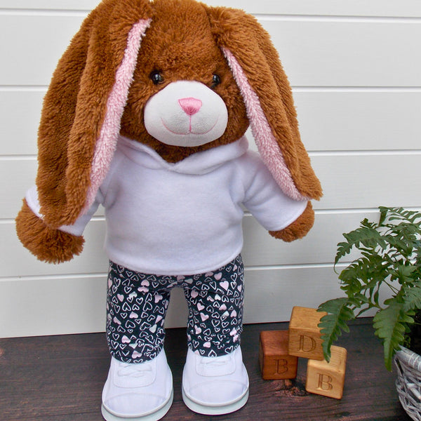 Build a bear teddy bear wearing a white hoodie, black and pink leggings and white training shoes. The teddy bear is standing in front of a white background with a green fern plant on the right. The teddy bear hoodie and teddy bear leggings have been made from sewing patterns by Best Dressed Bears.