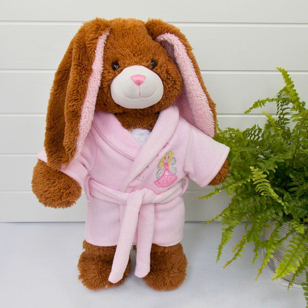 Brown teddy bear with long ears wearing a pink dressing gown with a fairy motif. The teddy bear is standing in front of a white background with a green fern plant to the right.