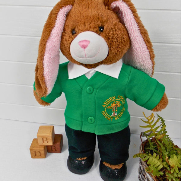 Brown teddy bear wearing a green school cardigan, a white polo t-shirt, black pants & black shoes. The teddy bear is standing in front of a white background with a green fern plant to the right and wooden bricks to the left. The teddy bear school cardigan, teddy bear t-shirt and teddy bear pants are all made from sewing patterns by Best Dressed Bears.