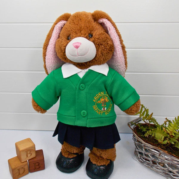 Brown teddy bear wearing a green school cardigan, a white polo t-shirt, a navy pleated skirt & black shoes. The teddy bear is standing in front of a white background. The teddy bear school cardigan, teddy bear t-shirt, teddy bear pants & teddy bear skirt are all are made from sewing patterns by Best Dressed Bears.
