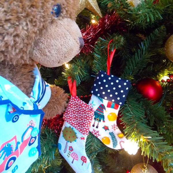 Teddy bear wearing blue patterned pyjamas standing in front of a Christmas tree. There are two mini Christmas stockings on the Christmas tree