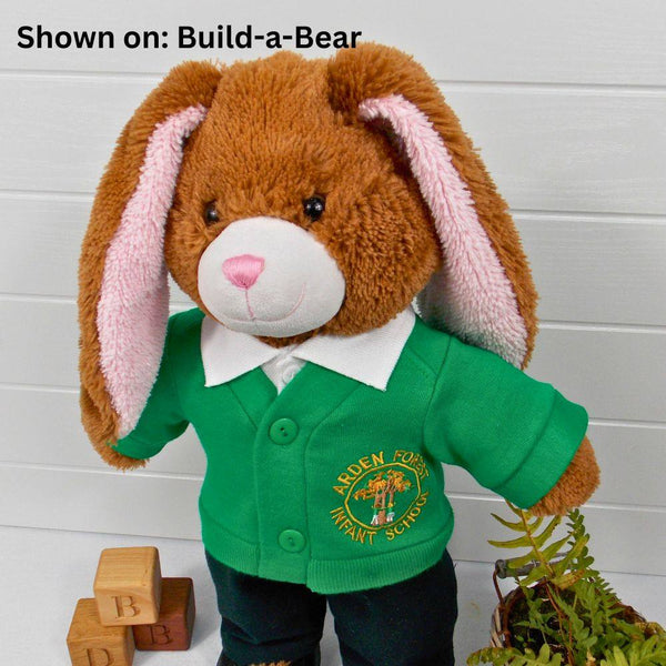 Build a bear teddy bear wearing a green school cardigan, white polo t-shirt & black trousers. The teddy bear is standing in front of a white background with a green fern plant to the right. The teddy bear school cardigan and teddy bear t-shirt are made from sewing patterns from Best Dressed Bears.