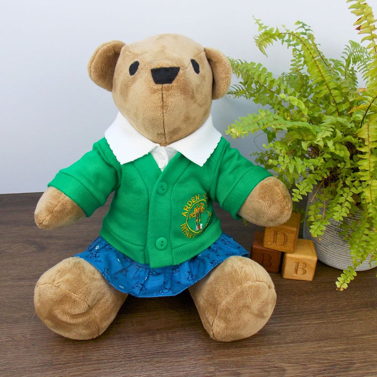 Funky Friends Factory Calico Bear wearing a green school cardigan, white polo t-shirt and blue patterned skirt. The bear is sitting on a wooden floor with a green fern plant to the right. The teddy bear clothes are made from sewing patterns by Best Dressed Bears